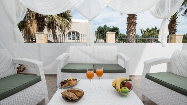 Un balcon agrémenté de meubles en osier blanc, une table avec des éléments pour le petit-déjeuner et une vue sur des palmiers et un immeuble.