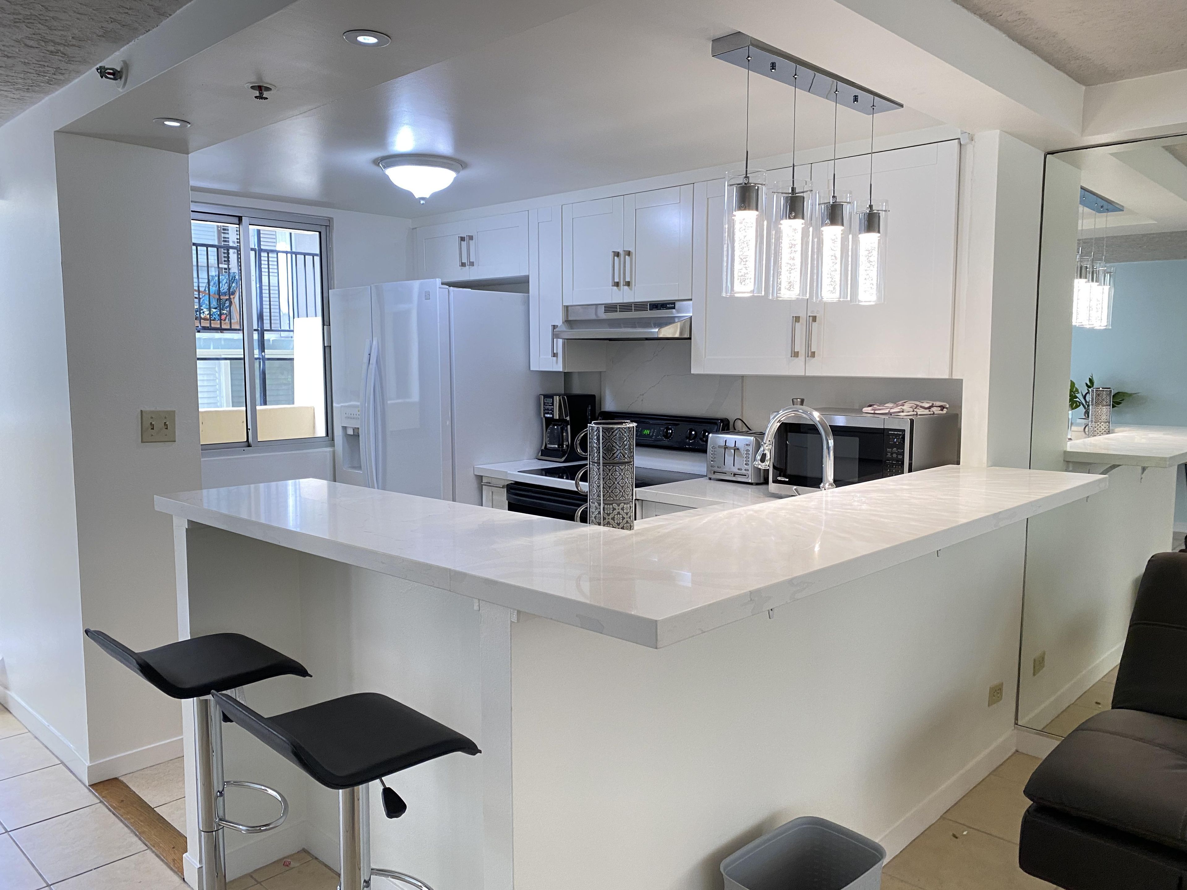 White quartz kitchen counter wraps around with two black bar stools beneath pendant lighting.