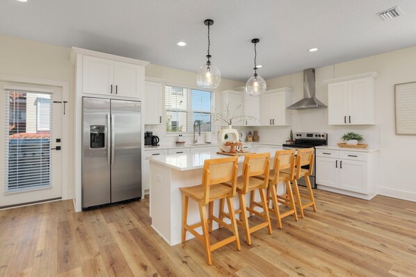 A modern kitchen with white cabinets, a stainless steel refrigerator, a central island with bar stools, pendant lighting, and a view of the outside through a window.