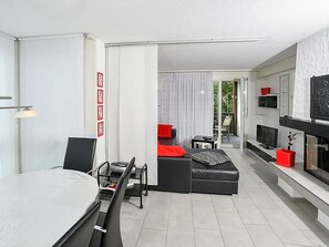 A modern living room with a dining area, a sofa with red cushions, a TV mounted on the wall, and a sliding glass door leading to a balcony.