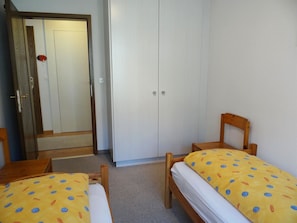 Ein Zimmer mit zwei hölzernen Betten mit gelber Bettwäsche, einem Holzstuhl, einem Kleiderschrank und einer Tür mit einem roten Gegenstand darauf.
