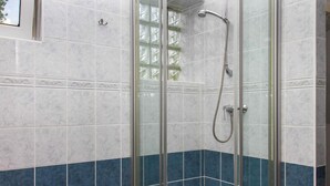Ducha con mampara de vidrio, cabezal de ducha de mano, azulejos blancos y azules en las paredes y un pequeño ventanal sobre la ducha.