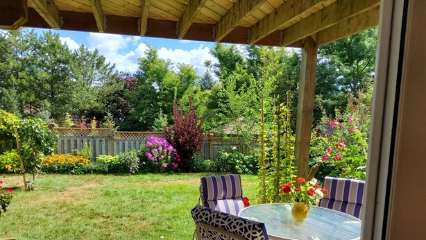 Eine überdachte Terrasse mit rundem Tisch und gestreiften Stühlen, umgeben von einem üppigen Garten mit verschiedenen Pflanzen und Blumen.