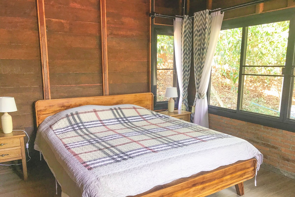 A wooden bed with a plaid blanket, a nightstand with a lamp, and a window with curtains.