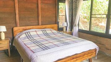 A wooden bed with a plaid blanket, a nightstand with a lamp, and a window with curtains.