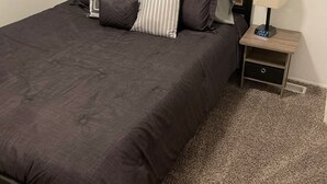 Un letto ben rifatto con una coperta scura e diversi cuscini, un comodino con una lampada e una sveglia digitale, e un pavimento coperto da moquette.