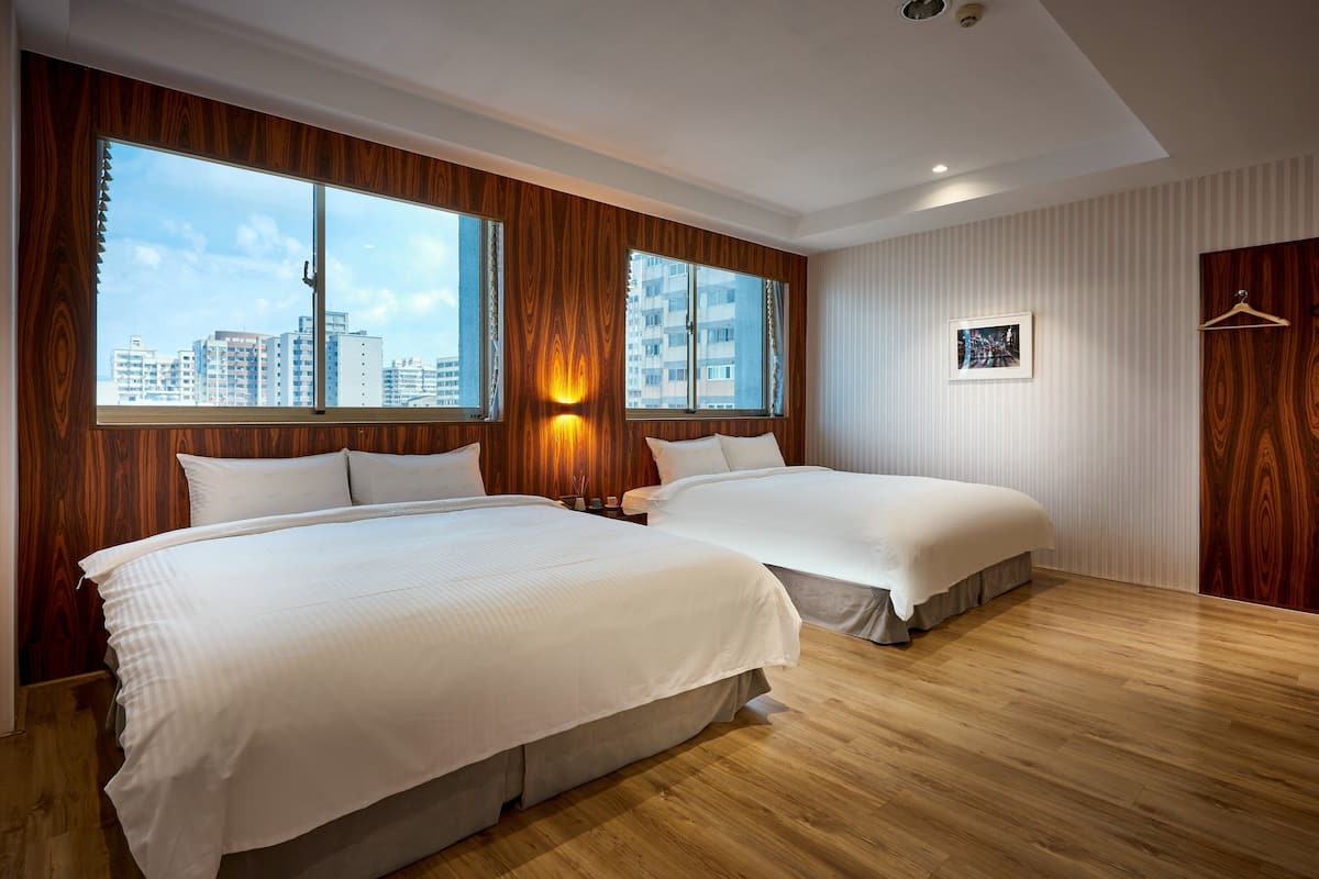 Hotellihuone, jossa on kaksi sänkyä, suuret ikkunat, näkymä kaupunkiin, puupanelointi ja seinällä oleva kehystetty taulu.