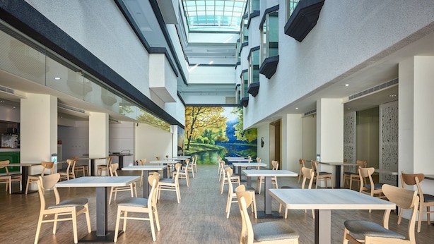 A modern indoor dining area with high ceilings, a skylight, and a mural of a landscape. Tables and chairs are arranged for dining. - Oursea Hotel (Taichung)