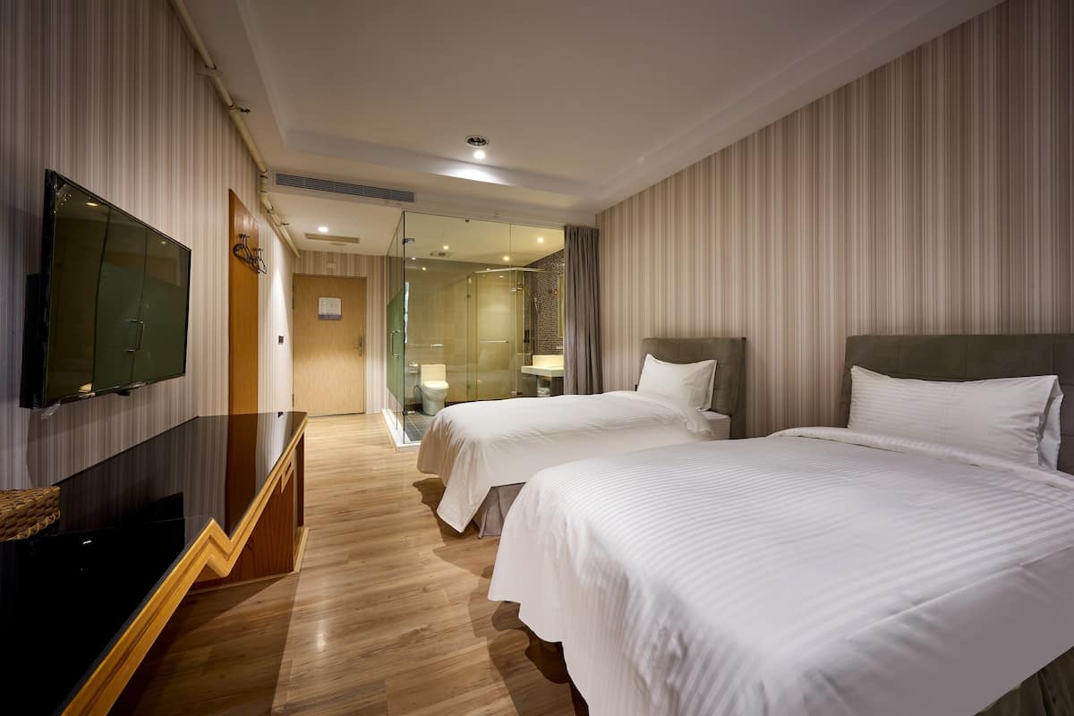 Hotellihuone, jossa on kaksi sänkyä, televisio, puinen portaikko ja kylpyhuone, joka näkyy lasiovesta.