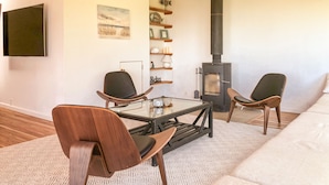 Ett modernt vardagsrum med ett soffbord i glas, två trästolar, en soffa, en öppen spis och en väggmonterad TV.