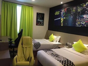 Ein Hotelzimmer mit zwei Betten, einem Schreibtisch, einem gelben Gaming-Stuhl und einem großen Fernseher, der ein Rennspiel zeigt.