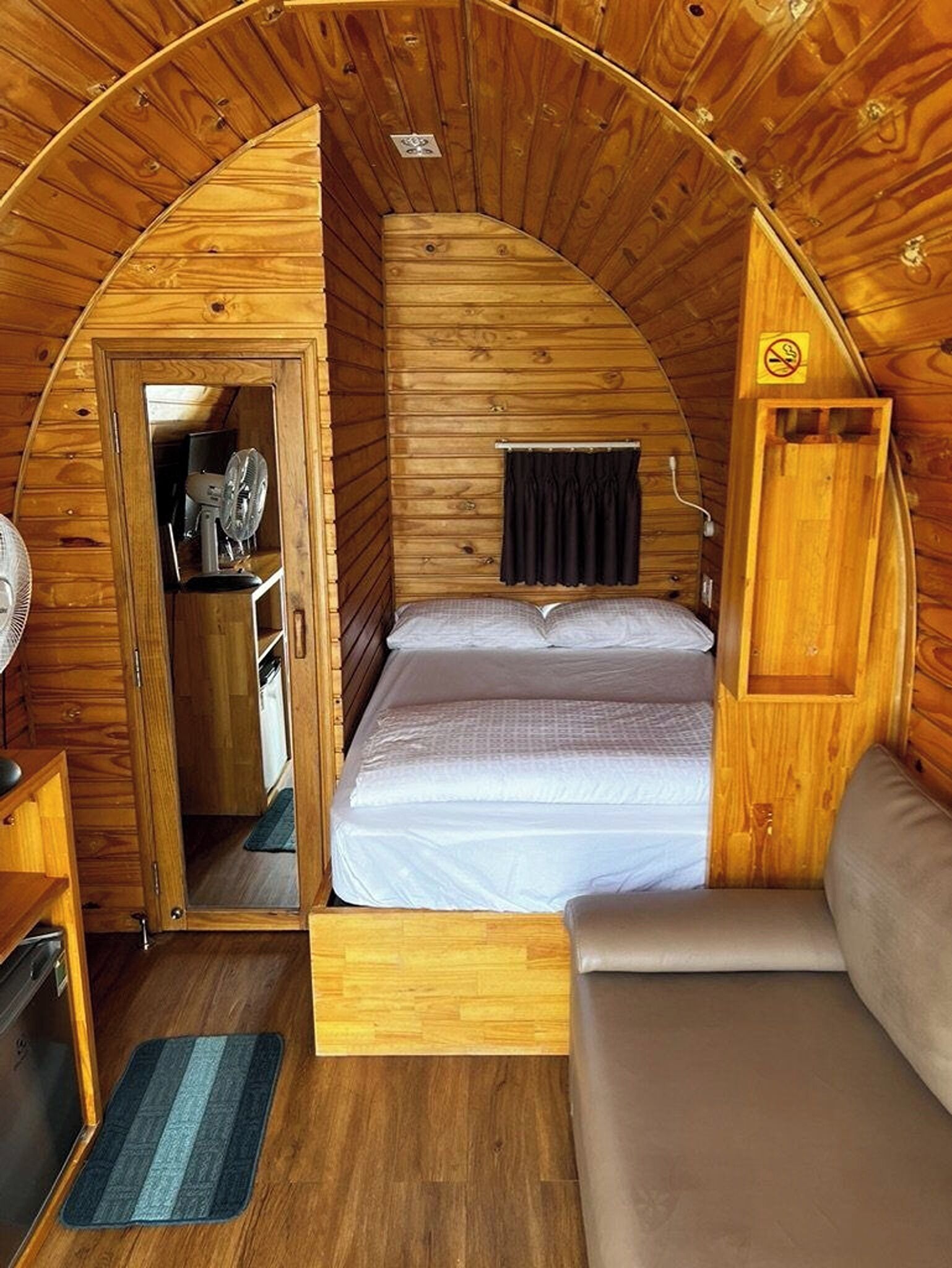 A cozy, wooden cabin interior with a bed, a small seating area, and a door leading to a kitchenette.