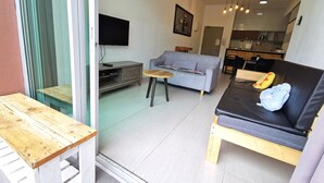 Ein modernes Wohnzimmer mit grauem Sofa, einem Holztisch und einem schwarzen Sofa mit blauem Kissen. An der Wand ist ein Fernseher angebracht und im Hintergrund befindet sich eine Küchenzeile.