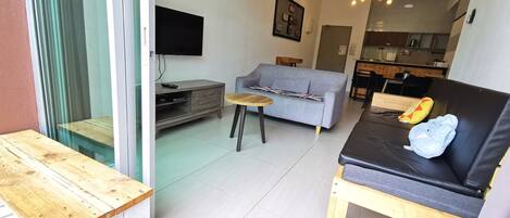 A modern living room with a grey sofa, a wooden coffee table, and a black sofa with a blue cushion. There is a TV mounted on the wall and a kitchen area in the background.
