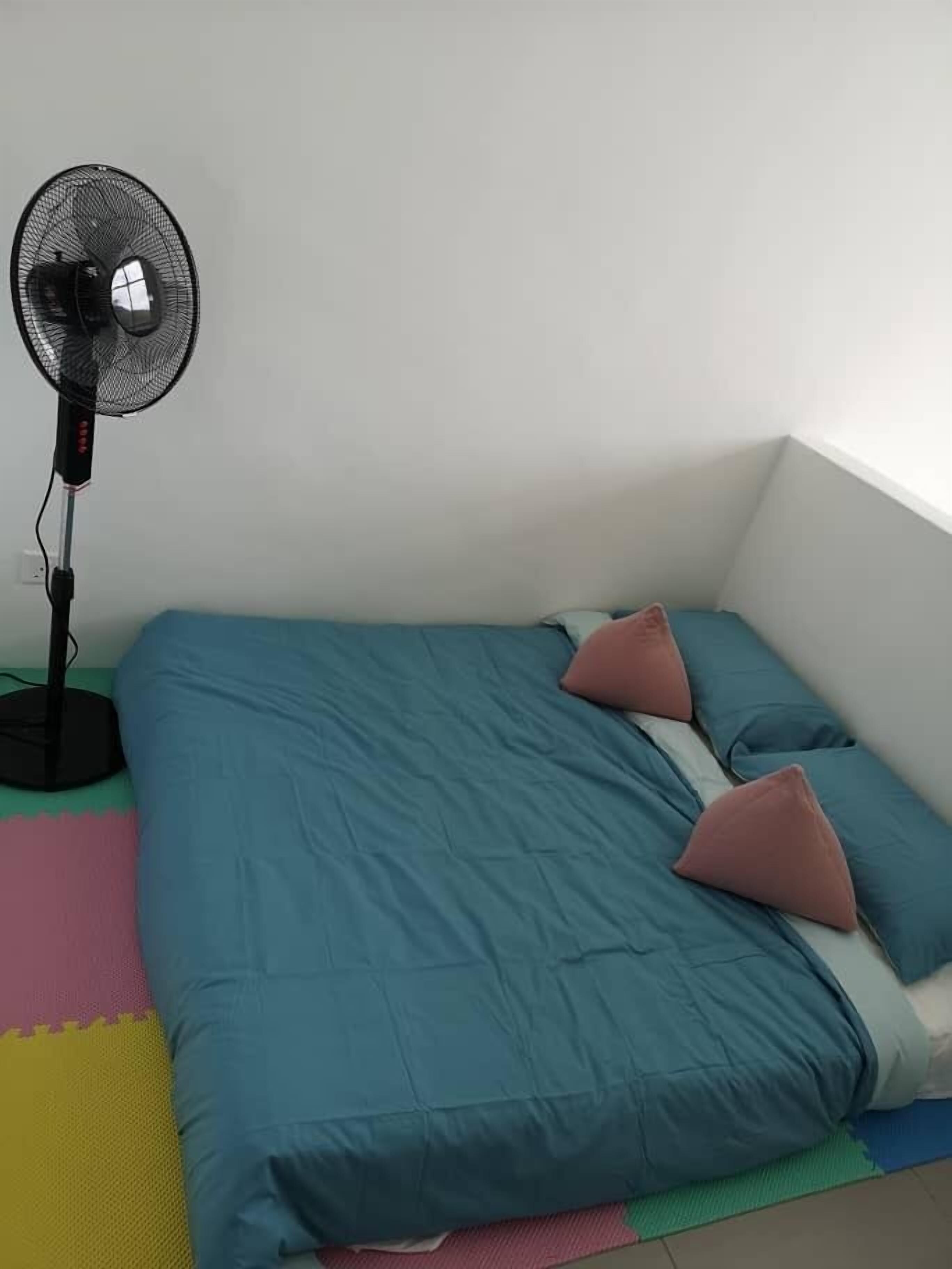 Une petite chambre avec un lit simple, une couvre-lit bleue, deux oreillers roses et un ventilateur sur pied.
