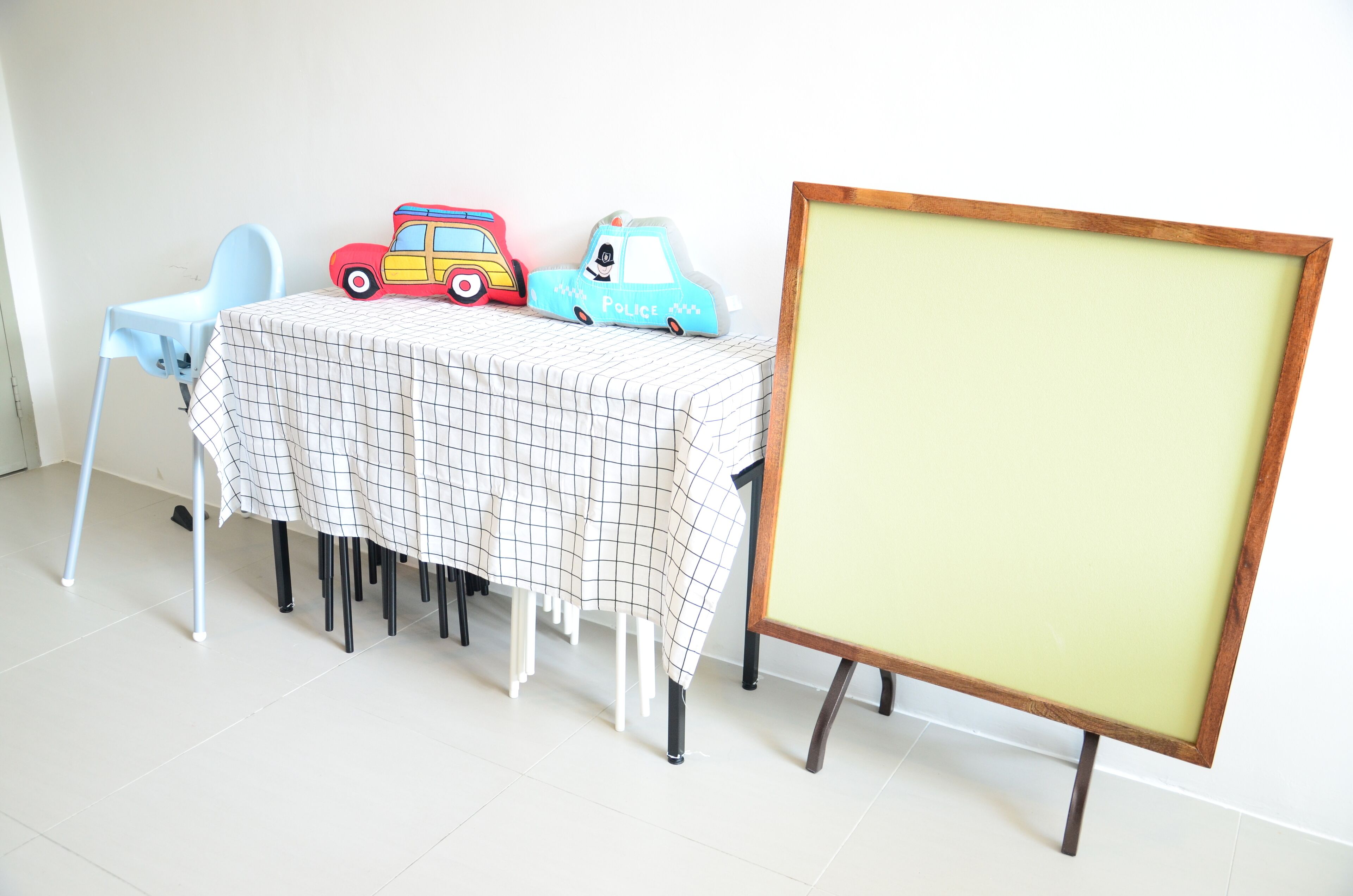 Ein Tisch mit karierter Tischdecke, zwei Spielzeug-Polizei-Autos, ein blauer Stuhl und eine leere gerahmte Tafel.