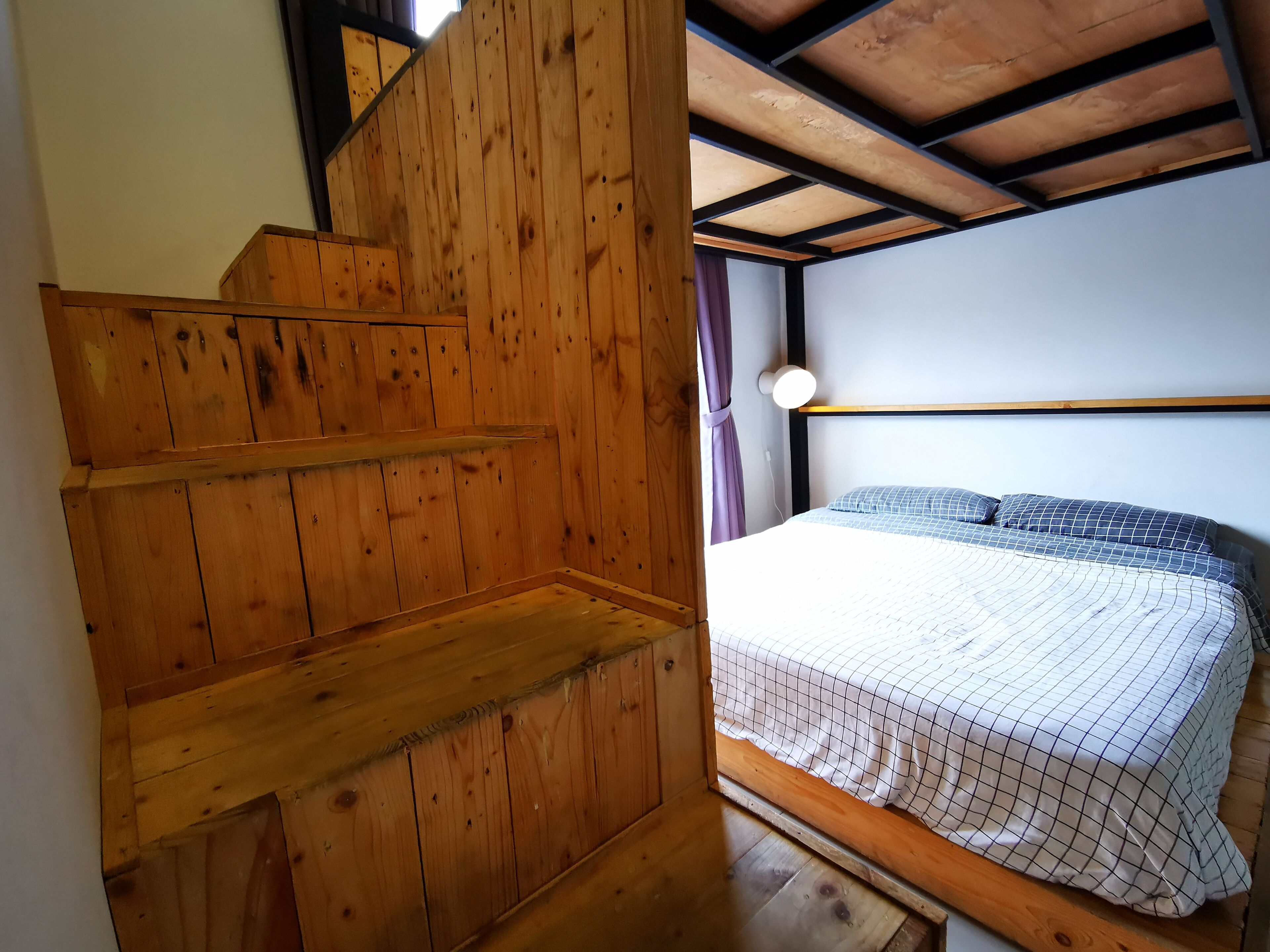 Uma escada de madeira leva a uma cama com cabeceira e janela com cortinas.