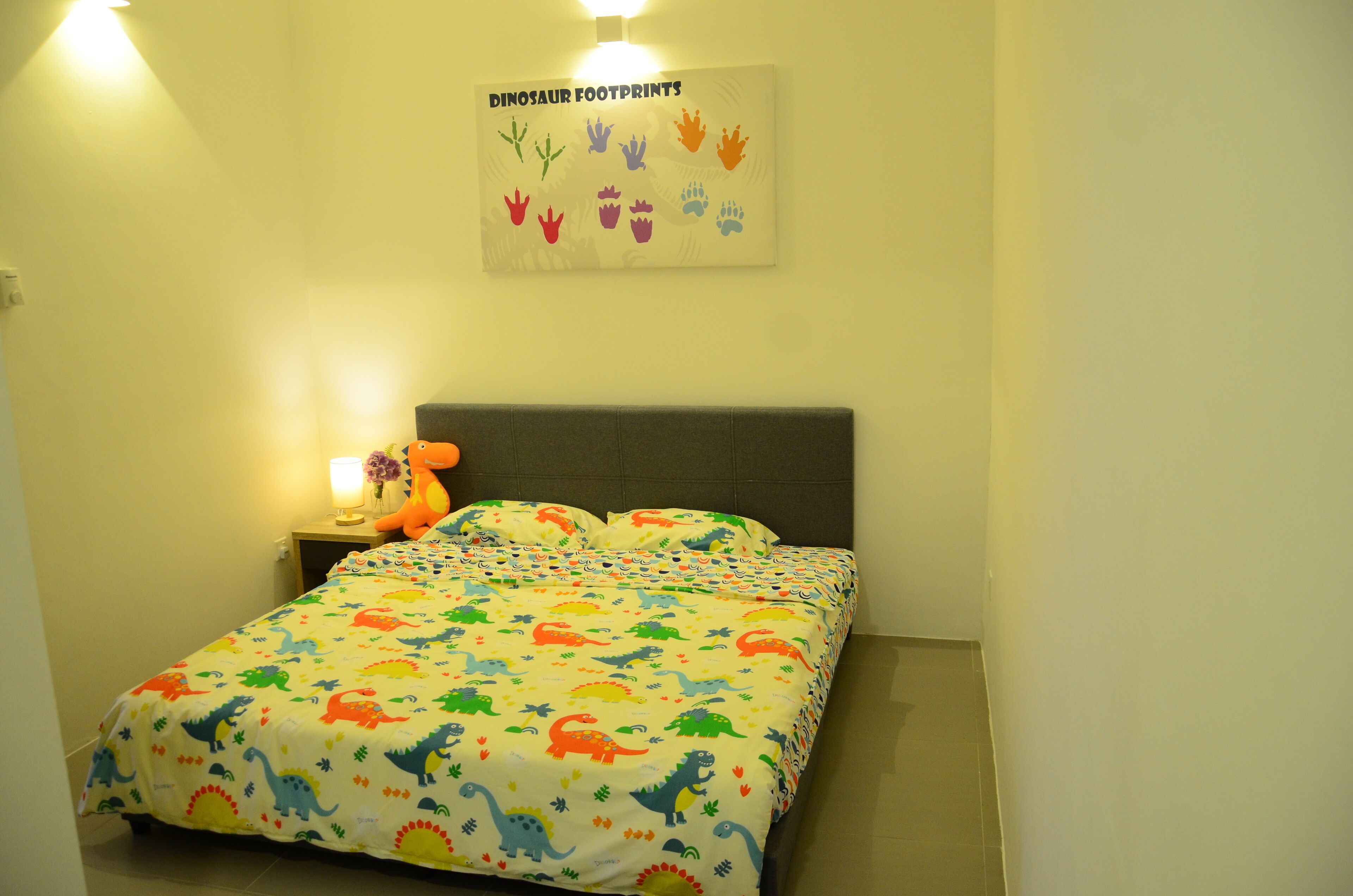 Une chambre à coucher avec un lit recouvert d’une couette à motifs de dinosaures, une table de chevet avec une lampe et un jouet en peluche, et un panneau décoratif fixé au mur au-dessus du lit.