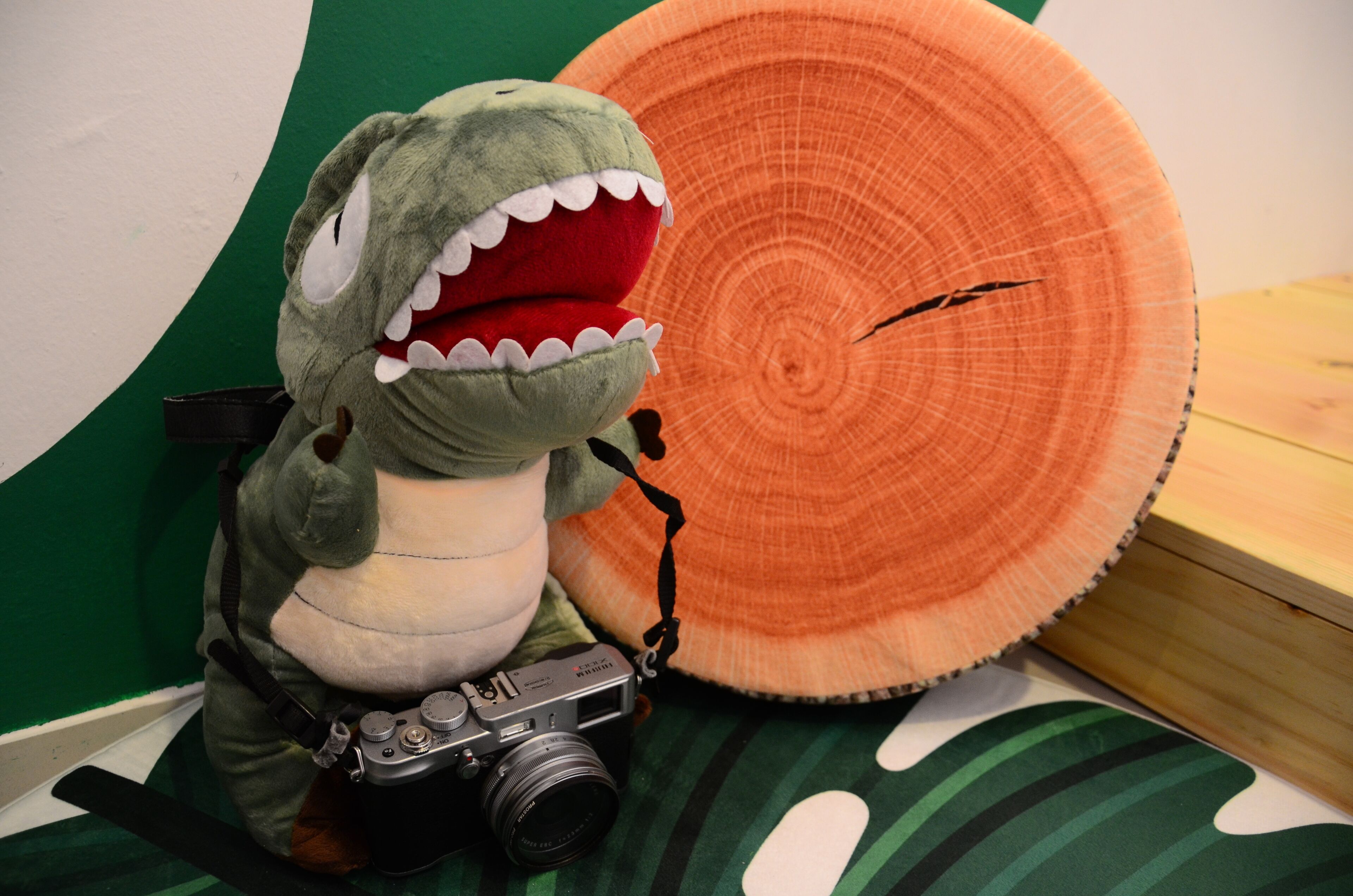 A plush toy dinosaur with a camera on a bed with a green and white striped pillow and a large orange circular object.