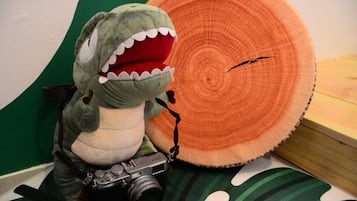 A plush toy dinosaur with a camera on a bed with a green and white striped pillow and a large orange circular object.