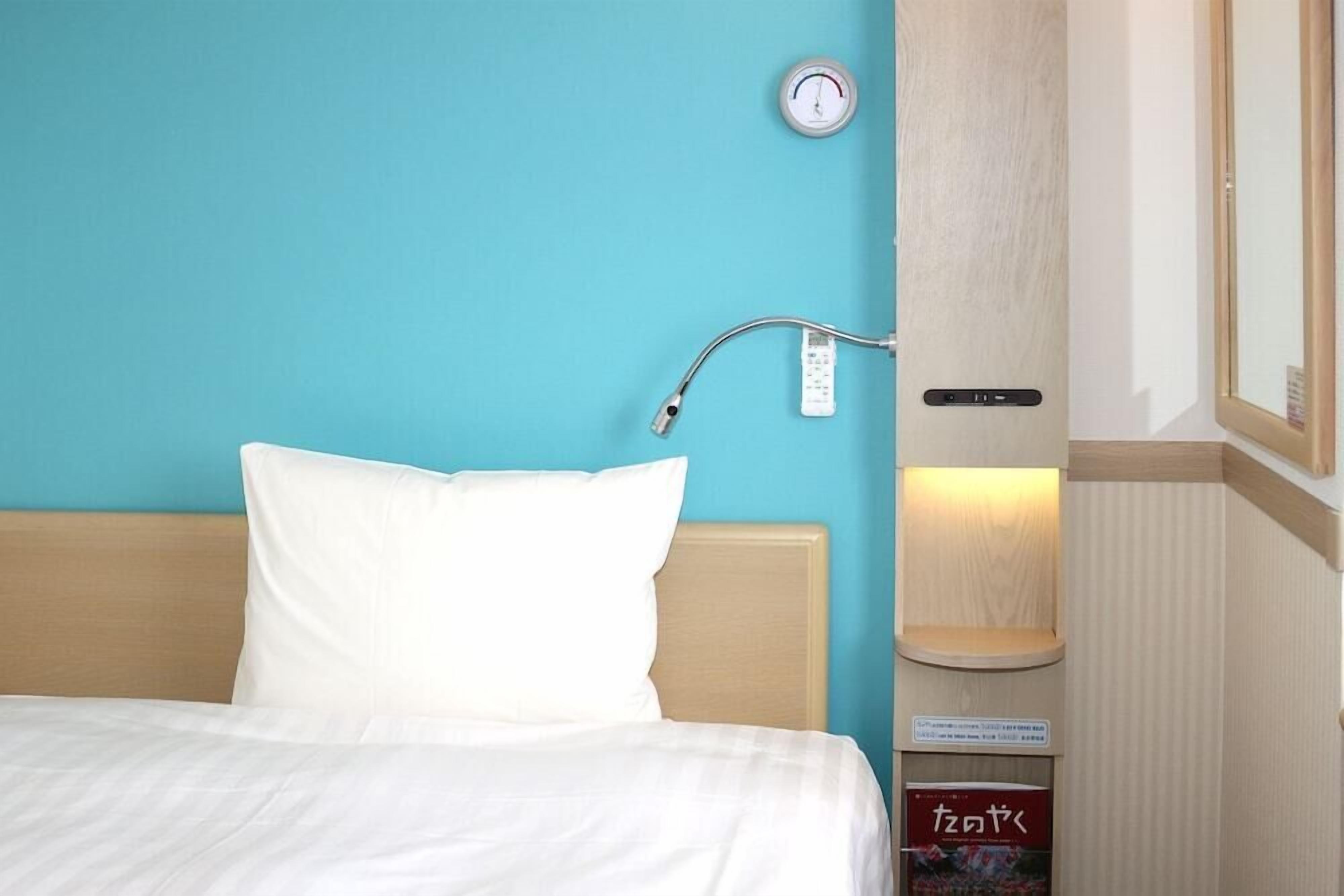 A hotel room with a bed, a nightstand with a lamp, a wall clock, and a small shelf with a book.