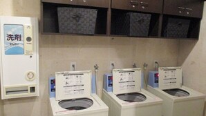 A laundry area with three washing machines, a detergent dispenser, and storage shelves above. - Toyoko Inn Akabane-eki Higashi-guchi (Tokyo)