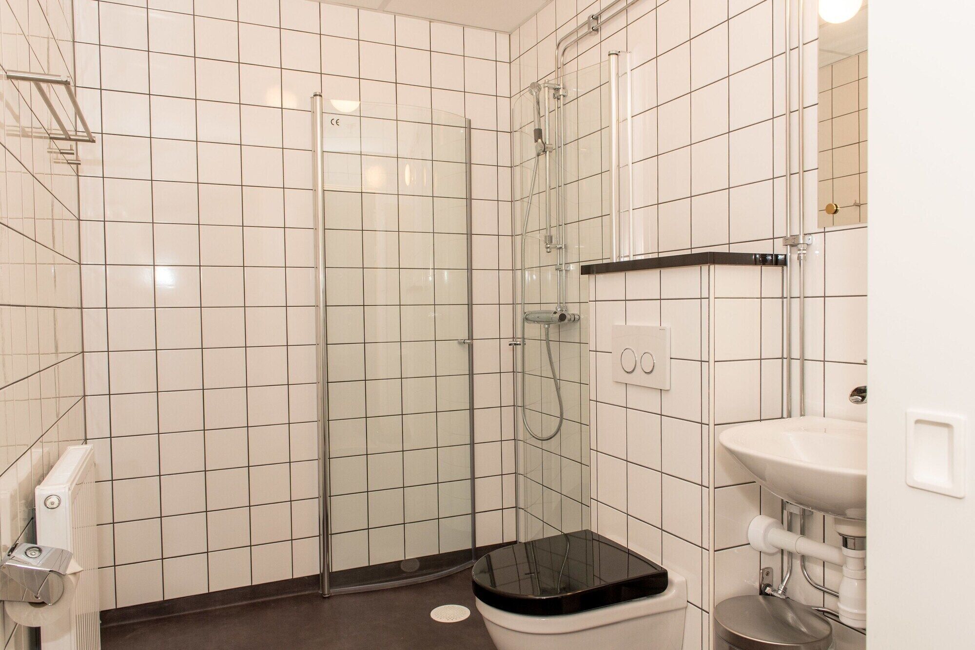 A modern bathroom with white tiled walls, a glass shower enclosure, a black toilet seat, and a white sink.