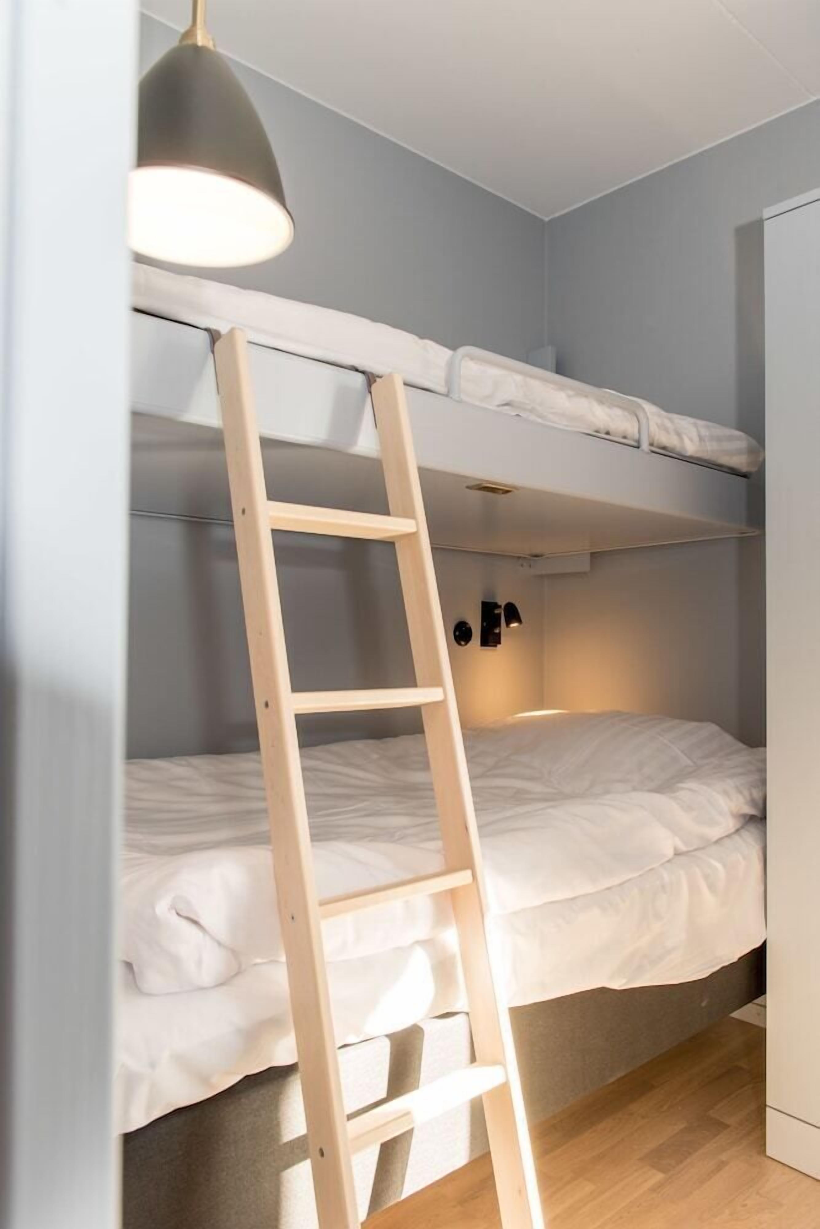 A bunk bed with a wooden ladder, a bedside table, and a wall-mounted light fixture.