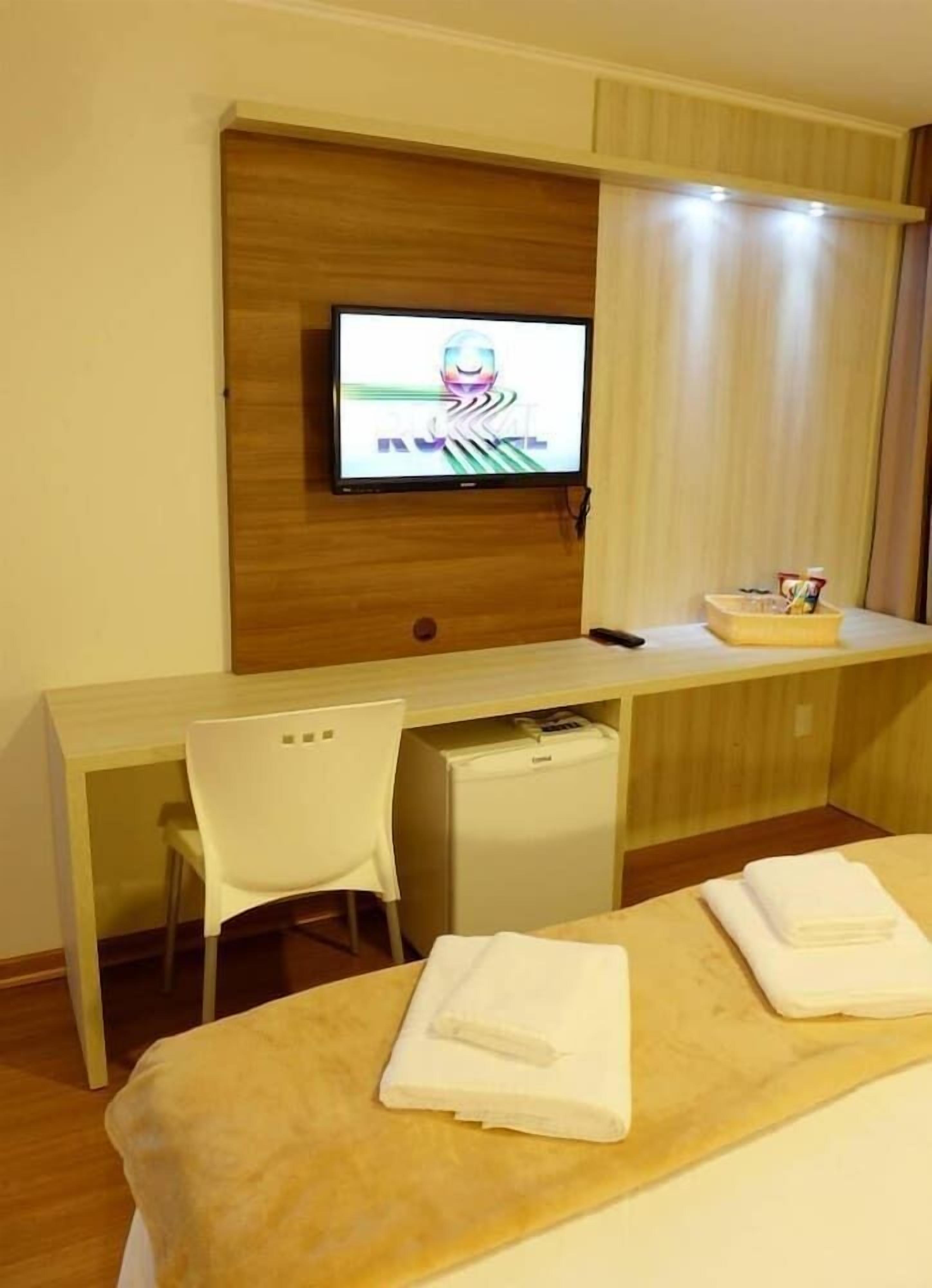 Habitación de hotel con un escritorio de madera, un televisor montado en la pared, un refrigerador pequeño, una silla blanca y una cama con toallas.