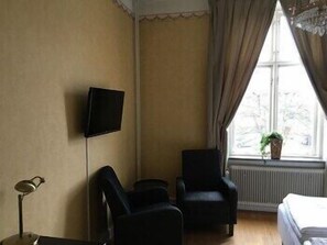 A room with a bed, two armchairs, a television on the wall, a radiator, and a window with curtains. - Stadshotellet i Sölvesborg (Sölvesborg)