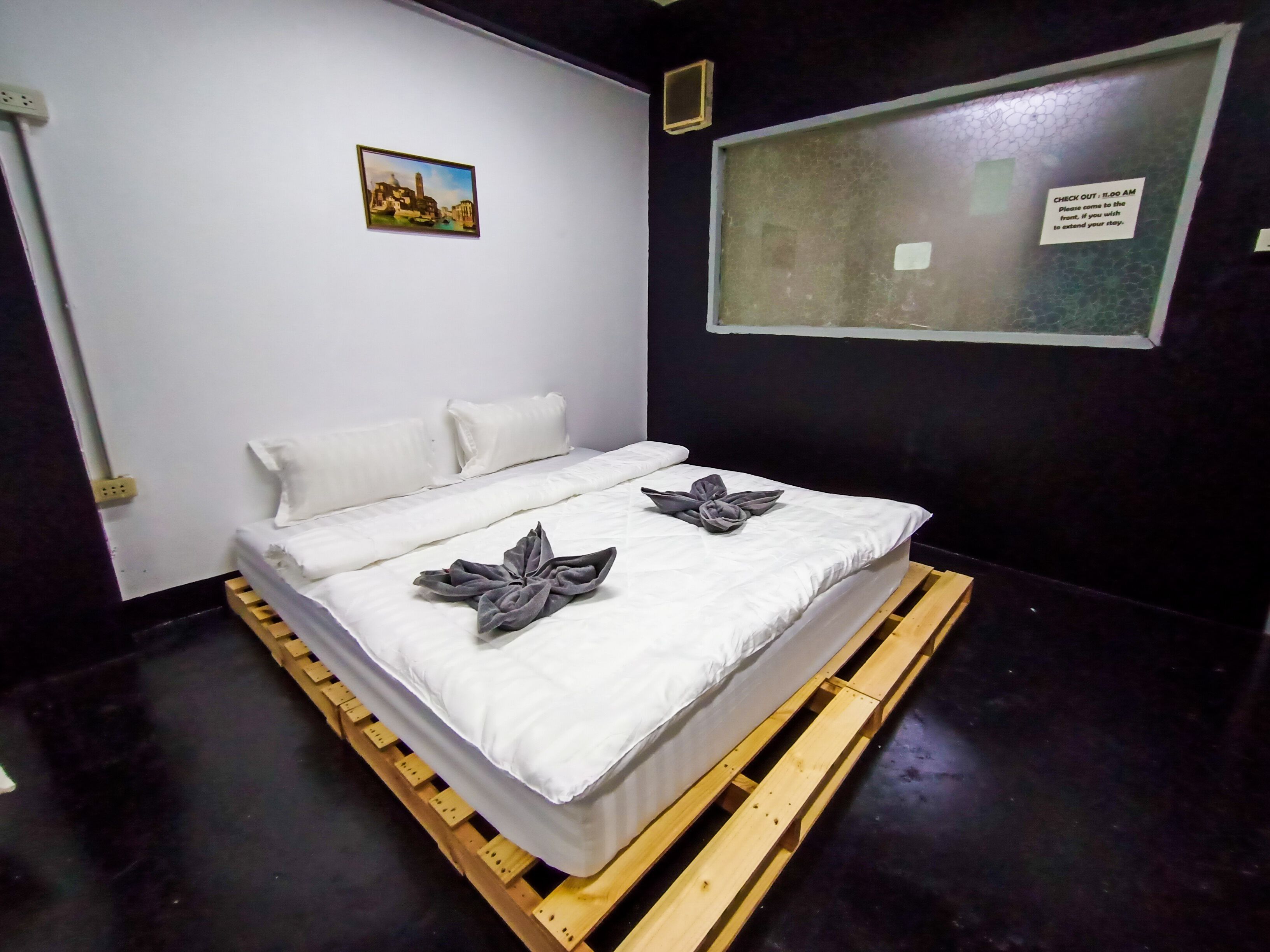 A hotel room with a bed, white bedding, a wooden pallet under the bed, a small picture on the wall, and a window with a check-out sign.
