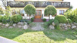 A two-story house with a wooden balcony, a stone pathway, and neatly trimmed bushes. - Apartment 1 with 2 bedrooms (4 adults + infant) on the ground floor - Villa Seeheim Ferienobjekt direkt (Boiensdorf)