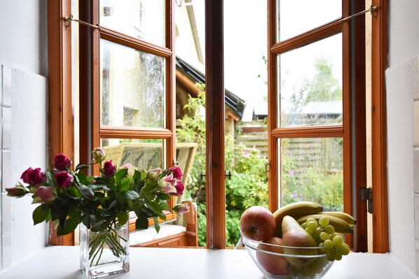 A window with a view of a garden, a vase with flowers, and a bowl of fruit on a windowsill.