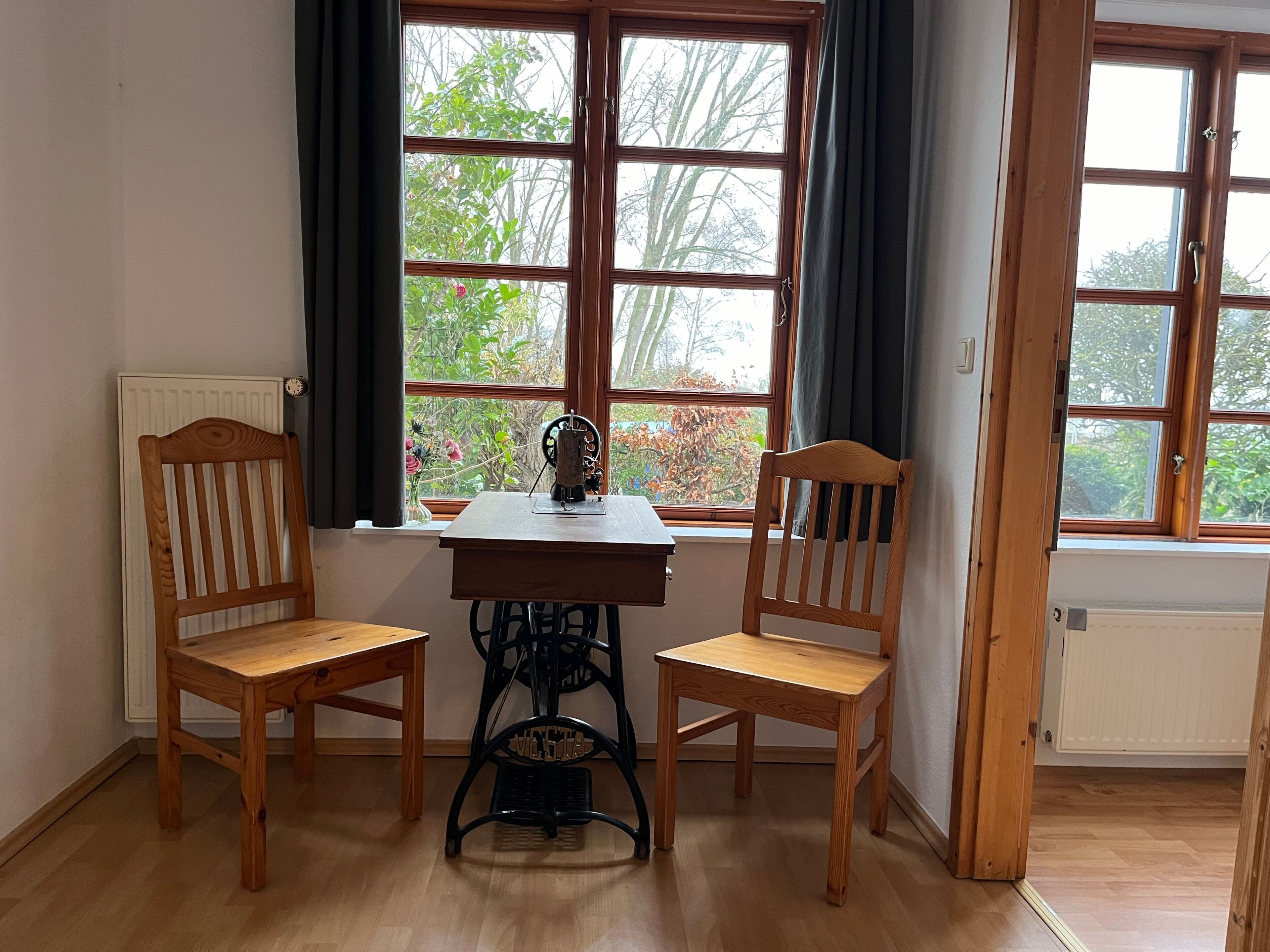 Ein Zimmer mit einem Holztisch, zwei Stühlen, einem Heizkörper und einem fenster mit Vorhängen.