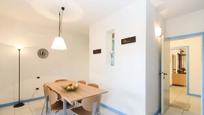 Een eetgedeelte met een houten tafel, vier stoelen, een fruitschaal, een vloerlamp en wanddecoratie.