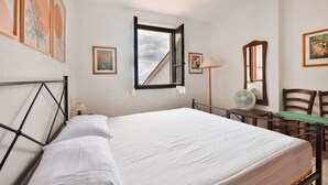A bedroom with a bed, a desk with a chair, a standing lamp, and framed pictures on the walls. - Cotoncello in a panoramic position (Marciana)