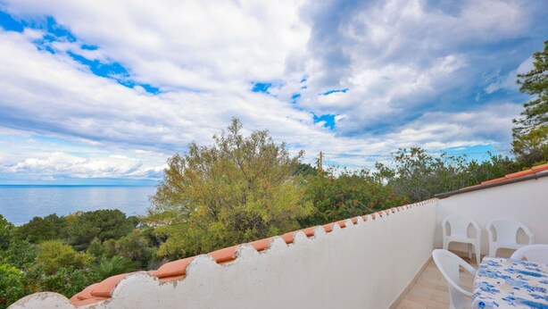 A balcony with a white wall, terracotta roof tiles, and a view of the sea. There are white plastic chairs and a table with a blue tablecloth. - Cotoncello in a panoramic position (Marciana)