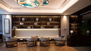 A modern lobby with a herringbone wood floor, a central seating area with sofas and armchairs, a dark bookshelf with decorative vases, and a ceiling with a circular light fixture.