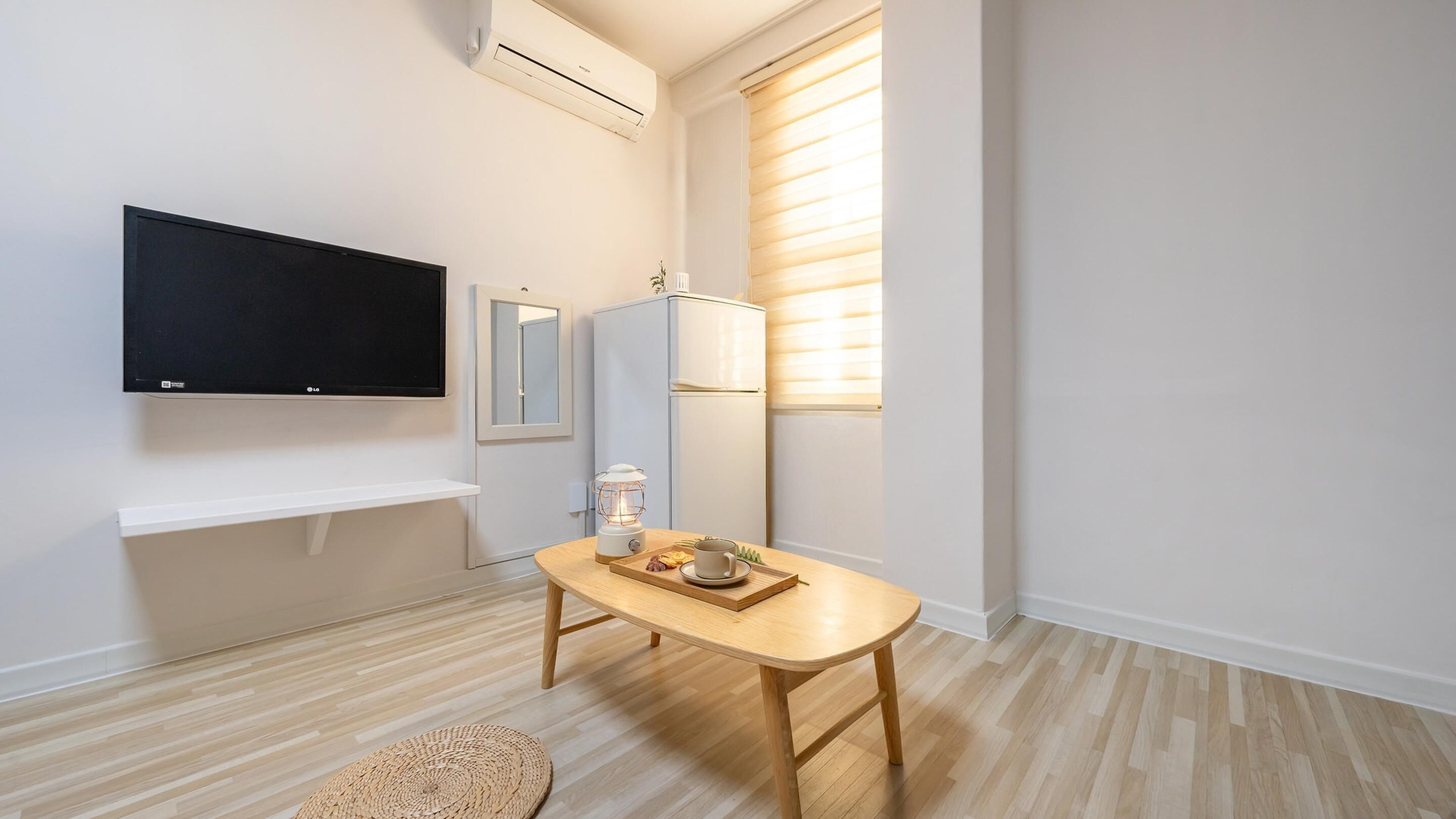 A modern, minimalistic living space with a flat-screen TV mounted on the wall, a small table with a cup and snacks, a compact refrigerator, and a window with blinds.