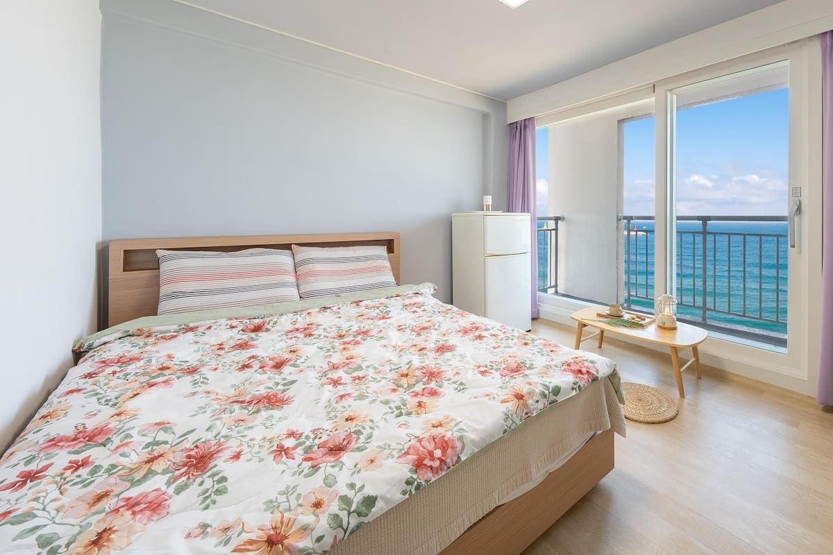Een slaapkamer met een bed, een klein tafeltje, een koelkast en een balkon met gordijnen.