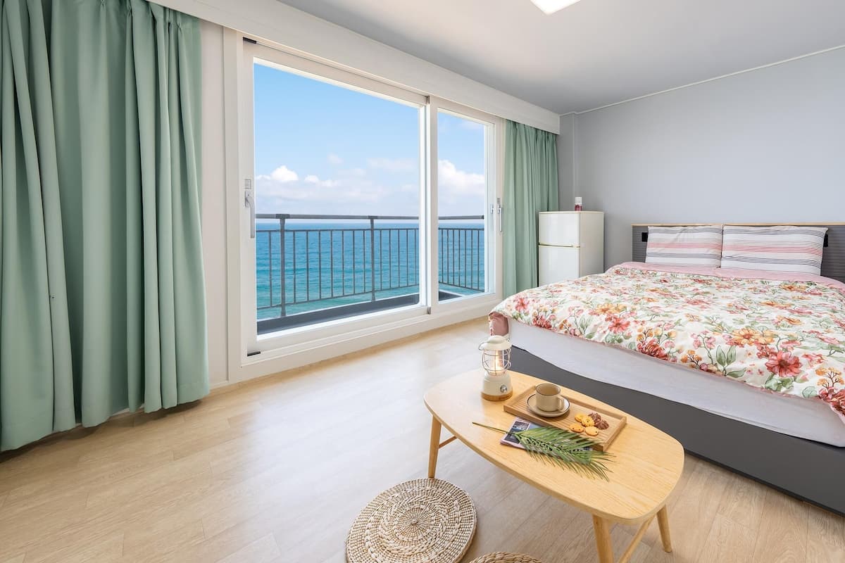 Een slaapkamer met een bed, een nachtkastje met een lamp, een raam met uitzicht op zee, en een klein tafeltje met een kopje en koekjes.
