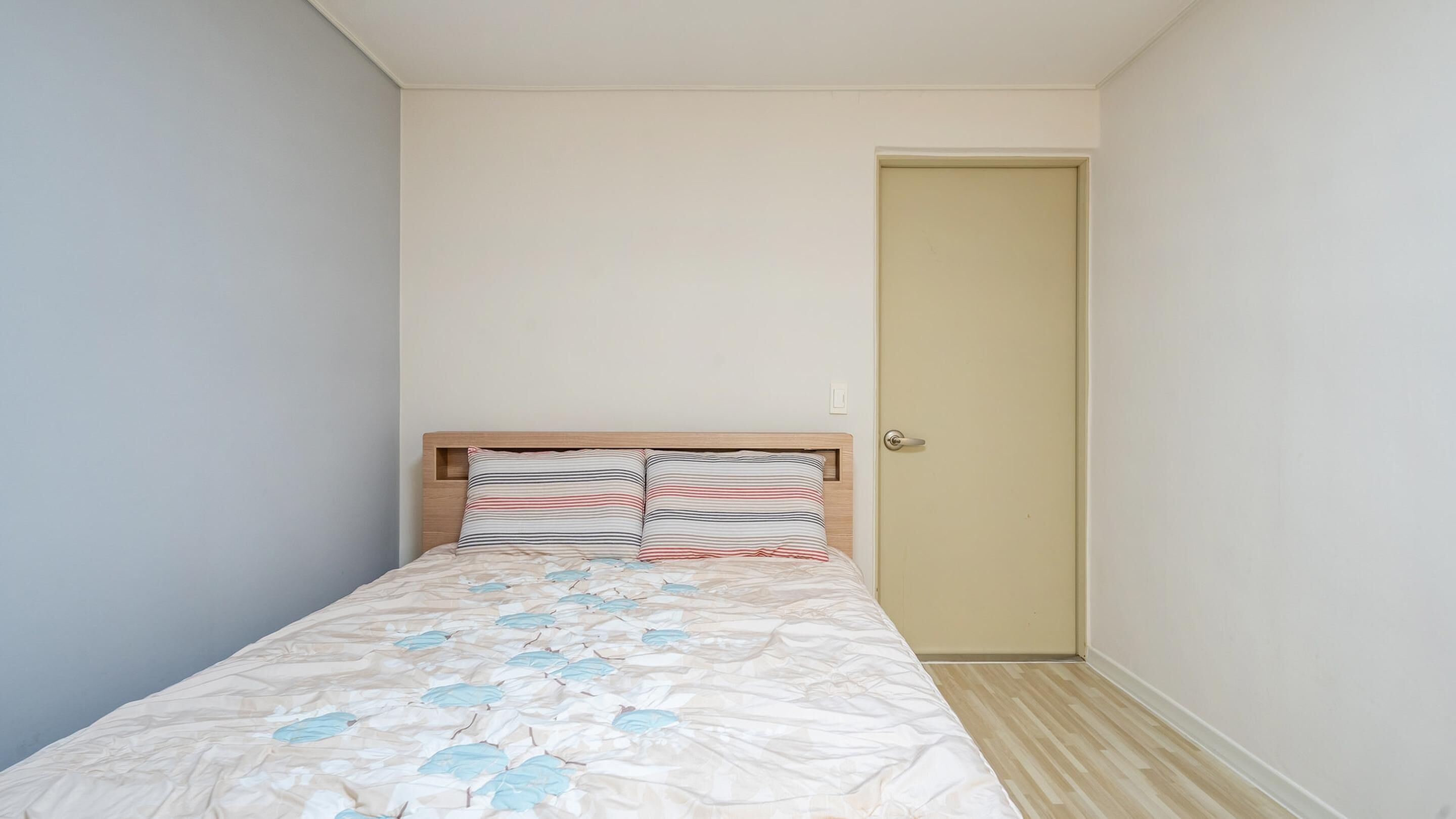 A small, single-bed room with a wooden headboard, a door, and a light-colored floor.