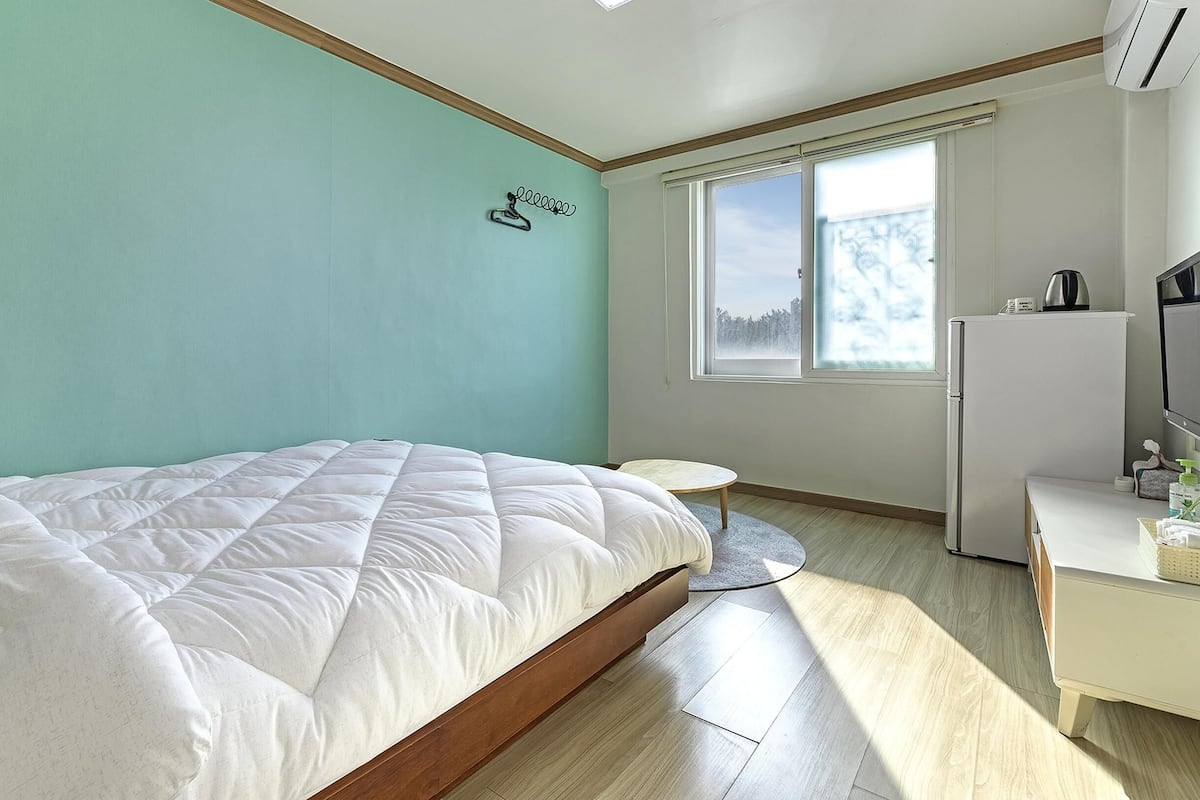 Een slaapkamer met een bed, een klein rond tafeltje, een witte dressoir en een raam met uitzicht op bomen.