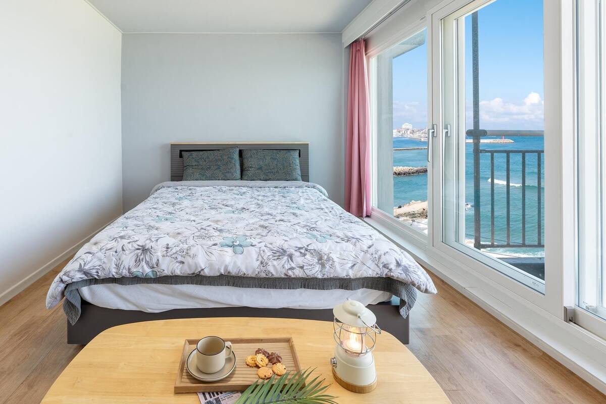 Een slaapkamer met een bed, een nachtkastje met een kopje en koekjes, een lamp en uitzicht op zee.