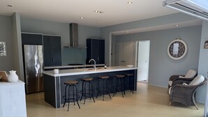 A modern kitchen with a central island, bar stools, a stainless steel refrigerator, and a built-in oven. - Entertainers Home at the Beach (Waihi Beach)