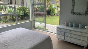 A bedroom with a bed, a dresser, and a large window with a view of a garden and a vehicle outside. - Entertainers Home at the Beach (Waihi Beach)