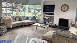 A living room with a sectional sofa, a flat-screen TV, a fireplace, and large windows with a view of greenery. - Entertainers Home at the Beach (Waihi Beach)