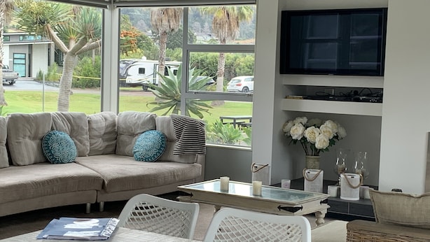 A modern living room with a sectional sofa, a glass coffee table, and a television mounted on the wall. - Entertainers Home at the Beach (Waihi Beach)