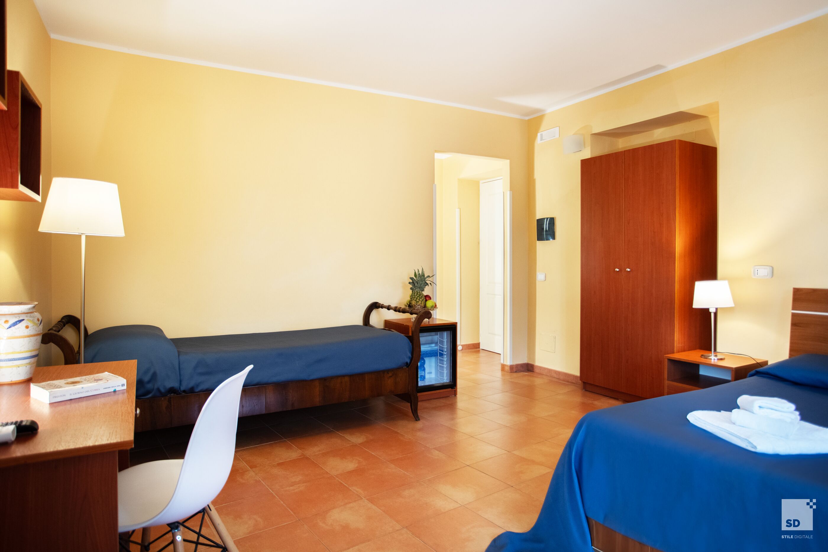 Ein Hotelzimmer mit zwei Betten, einem Schreibtisch mit Stuhl, einem kleinen Tisch mit Lampe und einem Kleiderschrank.