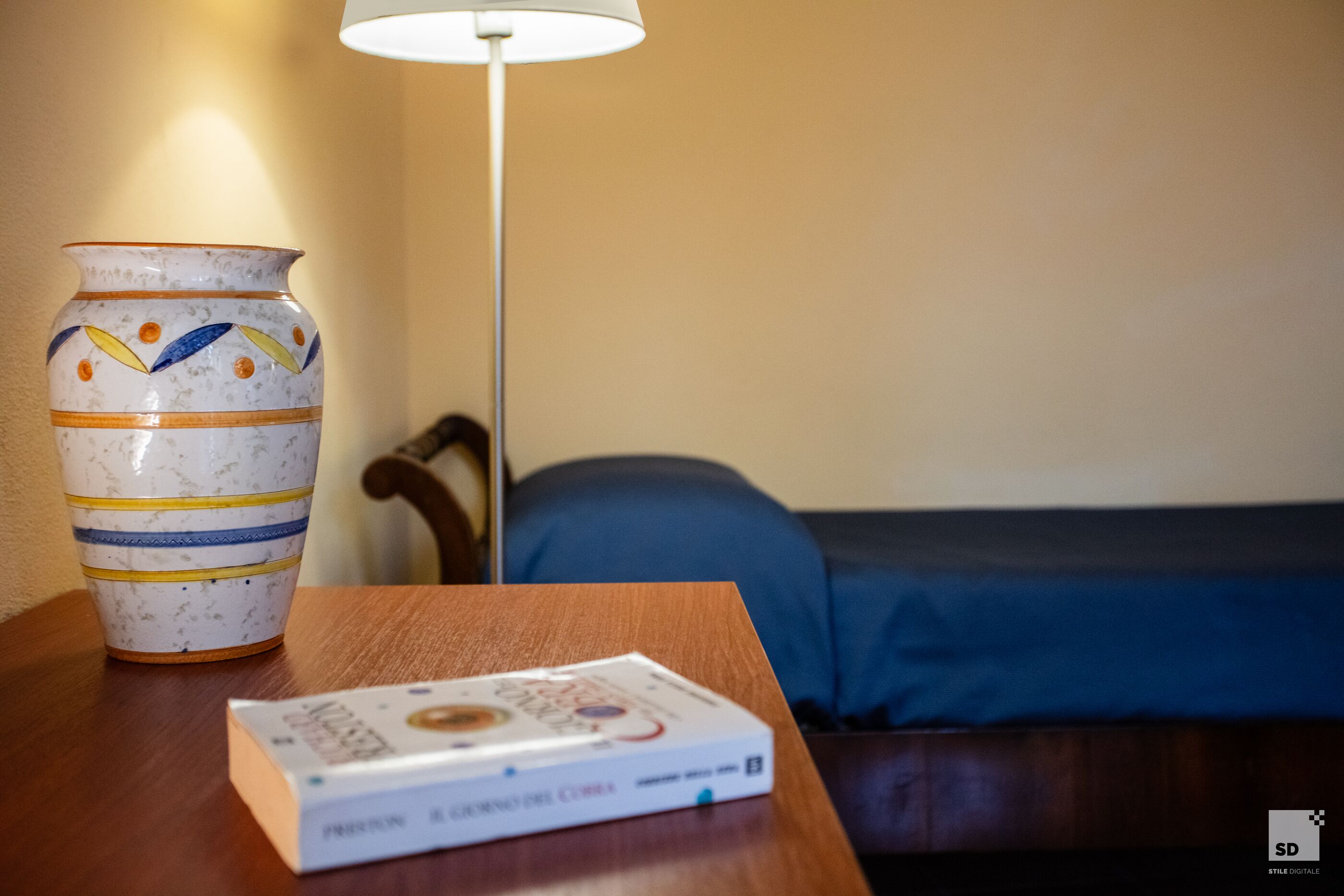 A room with a wooden table, a decorative vase, a book, a lamp, and a bed with a blue blanket.